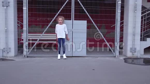 穿着白色毛衣和蓝色牛仔裤的卷发金发女孩正在一栋带有烤架的建筑背景上跳舞视频的预览图