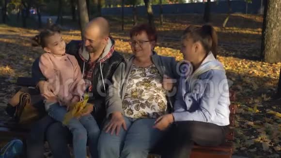 祖母带着两个孙子和成年儿子坐在秋天公园的长凳上视频的预览图