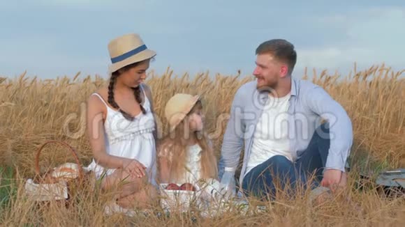 在户外一对幸福的夫妇和他们的小女儿坐在一起在秋天的金色谷物野餐中微笑着视频的预览图