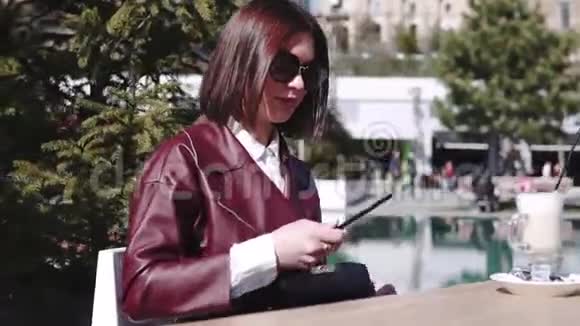 泳池附近咖啡馆里的女孩一位美丽的年轻女子在咖啡馆里喝着咖啡拿铁在阳光下使用她的智能手机视频的预览图