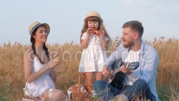 家庭户外假日年轻的爸爸弹吉他而他的妻子拍手他们的小女儿戴着草帽视频的预览图