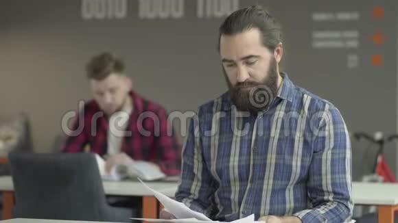 愤怒的胡子男坐在前台在办公室里研究文件工人们回头向他的同事扔东西视频的预览图