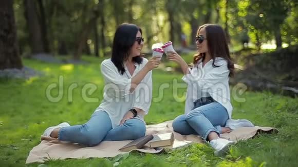 两个穿着白色衬衫的时髦快乐微笑的女孩正坐在公园里在柔和的绿色背景下谈笑风生视频的预览图