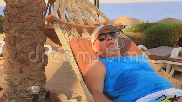 在埃及卡尔斯海的岸边一个戴着太阳帽的年轻人在一个阳光明媚的夏日在棕榈树的树荫下躺在吊床上视频的预览图