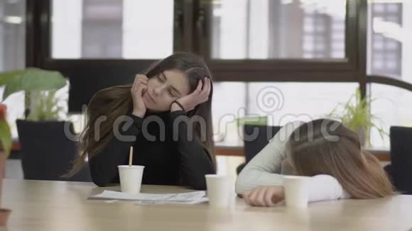 漂亮的女孩坐在桌子旁手伸在办公室里把咖啡混合在一起她的朋友靠在桌子上视频的预览图