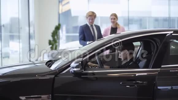 一对年轻成功的夫妇看着一个自信的女孩坐在驾驶座上一辆新的昂贵车的机舱里视频的预览图