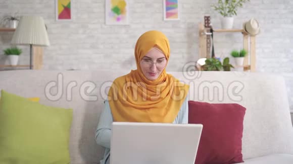 积极的年轻穆斯林妇女手提电脑坐在客厅的沙发上看着相机微笑使用银行卡视频的预览图
