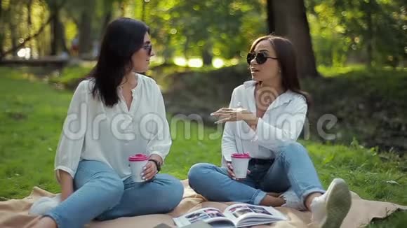 两个穿着白色衬衫的时髦快乐微笑的女孩正坐在公园里在柔和的绿色背景下谈笑风生视频的预览图
