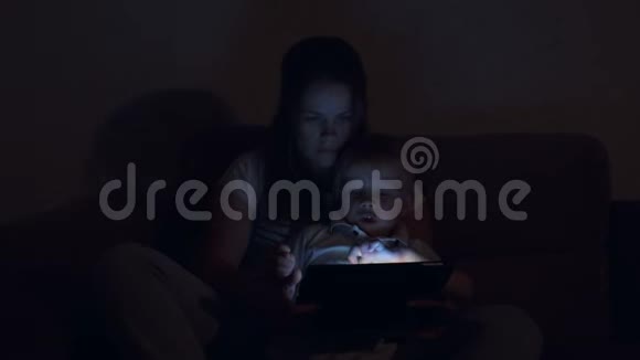 可爱的小男孩晚上在床上和妈妈在黑暗中玩平板电脑视频的预览图