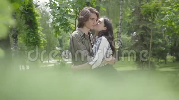 可爱的年轻夫妇温柔地拥抱和亲吻的背景模糊的叶子在前景快乐的女孩和男孩视频的预览图