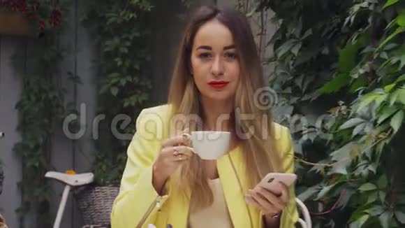 一个棕色长发和黄色外套的年轻漂亮女人坐在一家街头咖啡馆里微笑着喝着咖啡乌云密布视频的预览图