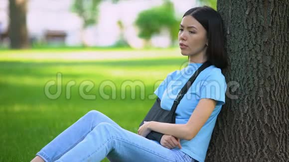 一位手臂受伤的年轻女士在公园里坐着一棵树药视频的预览图