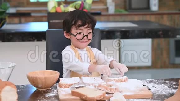 可爱的小亚洲男孩带着眼镜和围裙在家里的厨房里玩烘焙面包店作为厨师很有趣自制食物和面包视频的预览图