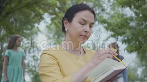 一位优雅的高级女性在前景公园里读这本书的肖像两个女孩躲在后面一个视频的预览图