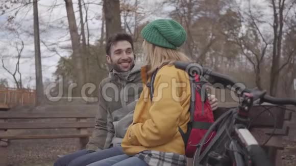 欢乐的一对游客带着背包坐在河岸的春天公园的长凳上有胡子的人和视频的预览图
