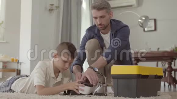 父亲和儿子在家地板上固定破碎的虚拟现实头盔特写孩子手里拿着百叶窗视频的预览图