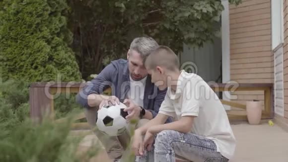 父亲和儿子坐在门廊上的肖像手里拿着一个泄气的足球特写家庭开支视频的预览图