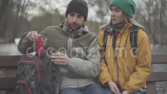 一对年轻的游客坐在春天公园的长凳上那个人从背包里拿出一个红色的热水瓶快乐视频的预览图