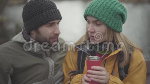 年轻夫妇穿着暖和的外套坐在户外说话的肖像穿着黄色夹克和绿色帽子喝酒的女人视频的预览图