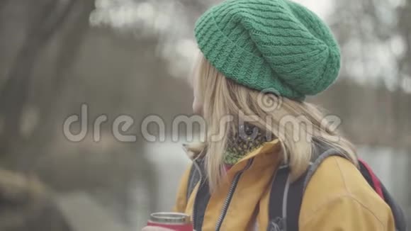 戴着绿帽子穿着黄色外套从热水瓶里喝茶或喝咖啡的自信美丽的年轻女子的肖像视频的预览图