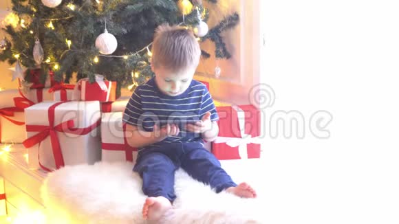 可爱的小男孩在圣诞树前用球和灯看动画片准备迎接新年的孩子视频的预览图