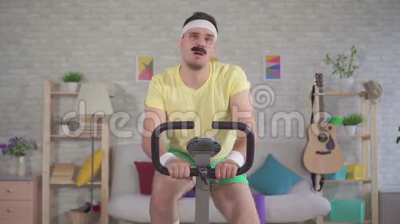 一位80的留着胡子的在家里骑着一辆静止自行车的滑稽精力充沛的男子运动员视频的预览图