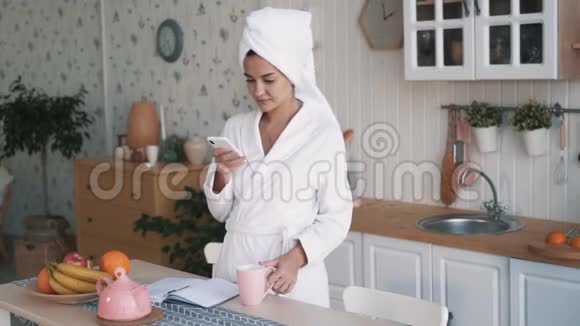 头上戴毛巾的女孩用电话在厨房喝咖啡慢动作视频的预览图