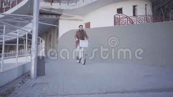 快乐微笑的人穿着棕色外套骑着自行车在大建筑附近的城市里英俊的城市居民的休闲视频的预览图