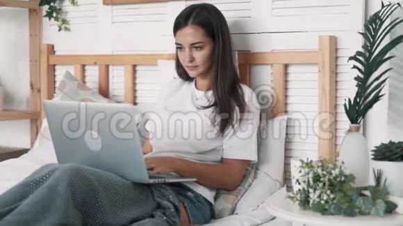 年轻女子坐在床上喝着咖啡在卧室的笔记本电脑上工作慢动作视频的预览图