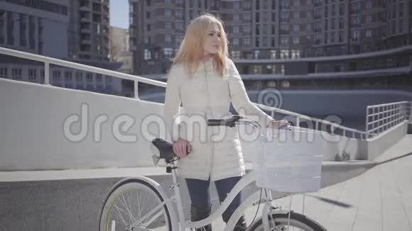 美丽的金发微笑的女人穿着温暖的白色夹克站在城市街道上骑着自行车看着镜头休闲视频的预览图
