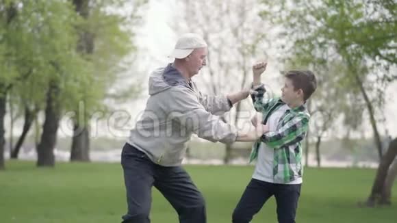 那个穿着格子衬衫的小男孩和积极的老人在公园里互相展示肌肉祖父和祖父视频的预览图