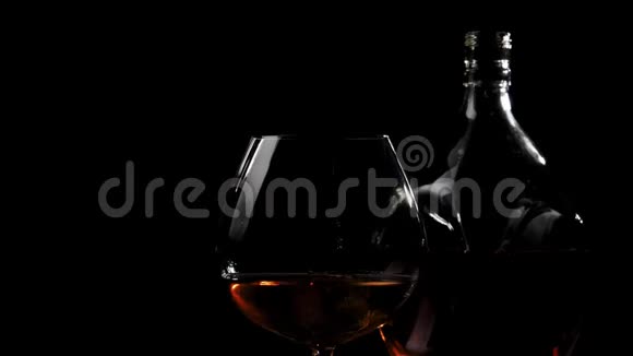 豪华白兰地手拿一个圆形瓶子与金色干邑从木桌附近的玻璃干邑在黑色背景视频的预览图