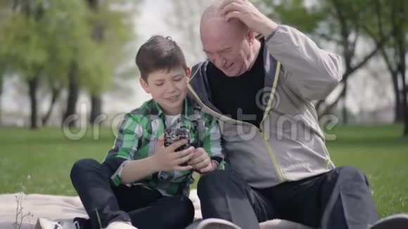 老人和孙子坐在公园的毯子上看着手机孩子教爷爷如何视频的预览图