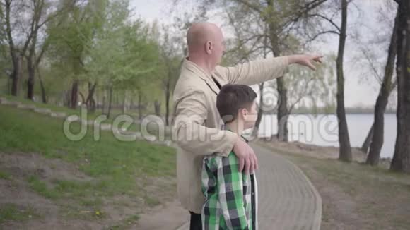 祖父和孙子站在河岸上望着水面指着远处那个人抱着孩子视频的预览图