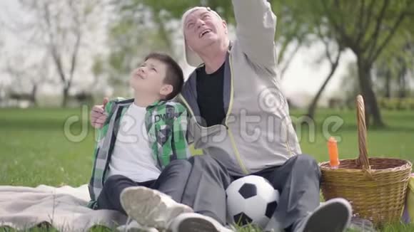 老人和孙子坐在公园的毯子上指着上面向男孩展示了一些细节家庭视频的预览图