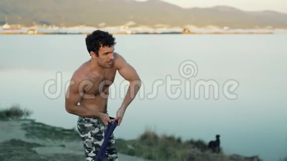 侧视肌肉男在山脉和大海的背景上用健身弹性橡皮筋做运动视频的预览图