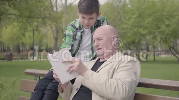 特写肖像可爱的祖父和可爱的孙子坐在公园的长凳上老人读着书视频的预览图