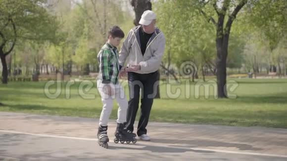 爷爷教孙子在公园里骑滑轮鞋老人牵着男孩的手帮助他视频的预览图