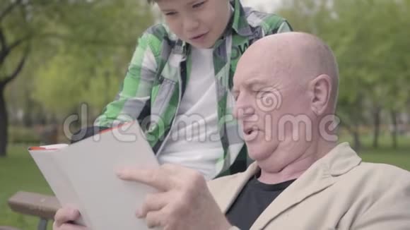 特写肖像可爱的祖父和可爱的孙子坐在公园的长凳上老人读着书视频的预览图