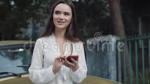 年轻迷人的笑容满面的黑发白种人女孩站在那里一边用手机一边靠在那辆时髦的金色旧车上视频的预览图