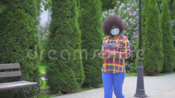 美国黑人女性留着非洲发型手里拿着智能手机戴着医用防护面具视频的预览图
