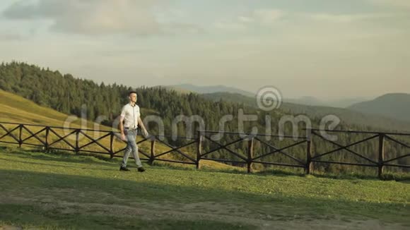年轻人站在山附近的田野里在他身后吃草的马日落年轻人走在整齐的篱笆上视频的预览图