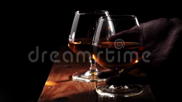 豪华白兰地在黑色背景的棕色木桌上放两个带有金色干邑的玻璃杯手拿右杯喝视频的预览图