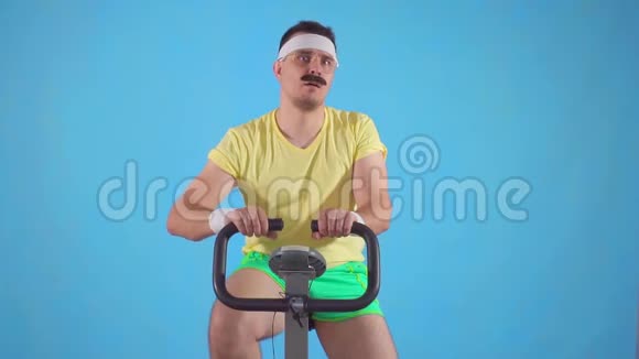 80岁留胡子的年轻滑稽男子的肖像手指朝下坐在运动自行车上慢动作视频的预览图