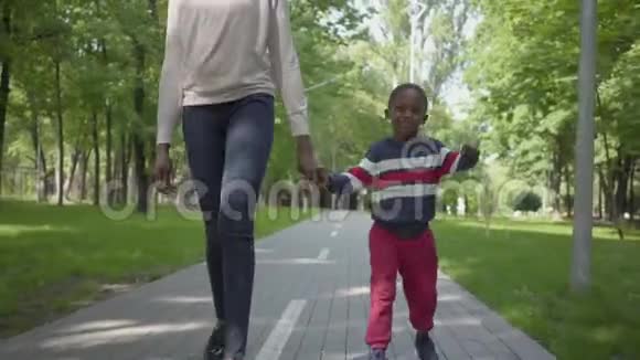 无法辨认的非裔美国妇女和她的小儿子牵着手在公园里散步可爱的孩子花时间视频的预览图