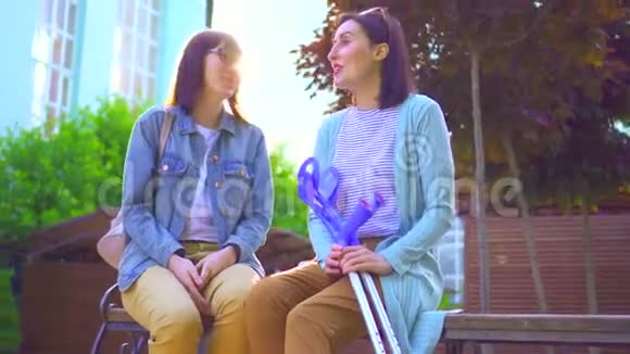 积极的年轻女子受伤拄着拐杖坐在公园的长凳上和一个朋友一起玩视频的预览图