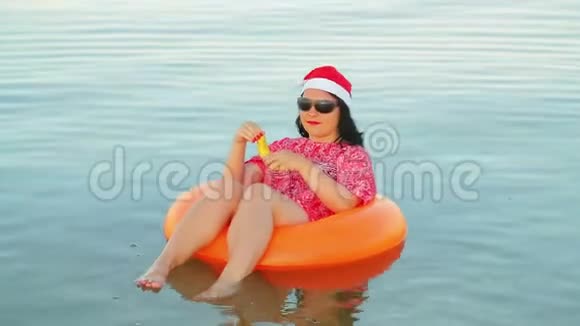 一个游泳圈里的黑发女人和一顶圣诞老人的帽子在海上吃香蕉庆祝圣诞节和新年视频的预览图