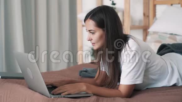 女孩的肖像躺在床上肚子上类型在笔记本电脑键盘上慢动作视频的预览图