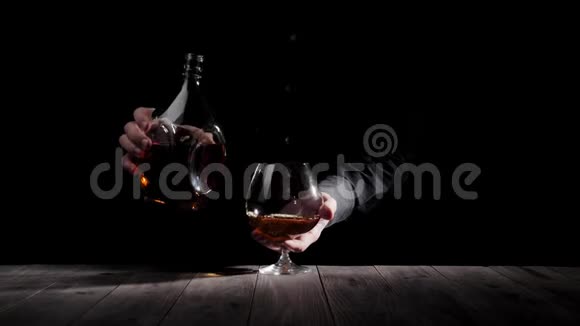 豪华白兰地穿着黑色衬衫的人把一个圆形瓶子放在靠近玻璃的木桌上背景是黑色的干邑视频的预览图