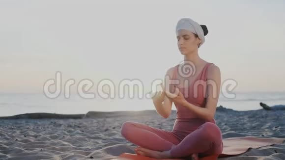 美丽健康的女性在日落时分坐在海滩上的莲花姿势练习瑜伽举起双手飞向天空呼吸视频的预览图
